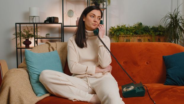 Bored tired young woman at home apartment talking on wired vintage telephone of 80s, fooling, making silly faces. Adult sleepy girl having unpleasant conversation in daytime living room on couch