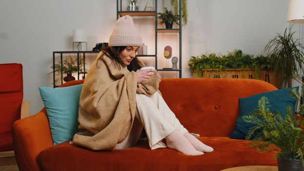 Sick young woman wrapped in plaid sitting, shivering from cold on sofa drinking hot tea in unheated apartment without heating due debt. Unhealthy girl in hat feeling discomfort try to warm up at home