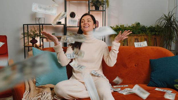 Money cash dollar rain falling on young woman celebrating success, winning, goal achievement, good news, lottery jackpot luck, dream come true at home apartment indoors. Career grow. Girl sits on sofa