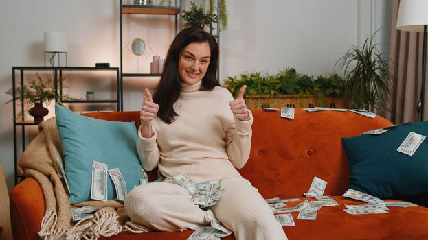 Successful happy rich business woman counting money cheering up with high profits, lottery game win, satisfied of wealth income earnings salary. Girl at home room couch full of dollar cash banknotes