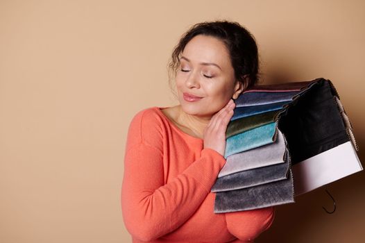 Middle-aged pretty woman in bright orange dress, touching upholstery materials in the fabric swatches, testing its texture for softness, posing with her eyes closed, isolated on beige color background