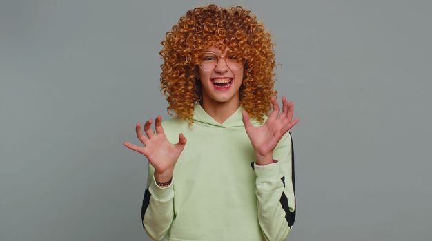 Funny furious teenager girl with curly frizzy hairstyle pretending to be a tiger, imitate parody of lion fierce shout roar, making cats claws scratching moves, flirting. Child kid on gray background