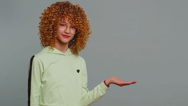 Happy teenager girl showing thumbs up pointing empty place, advertising area for commercial text, copy space for goods promotion banner, copy-space. Young child kid with curly hair on gray background