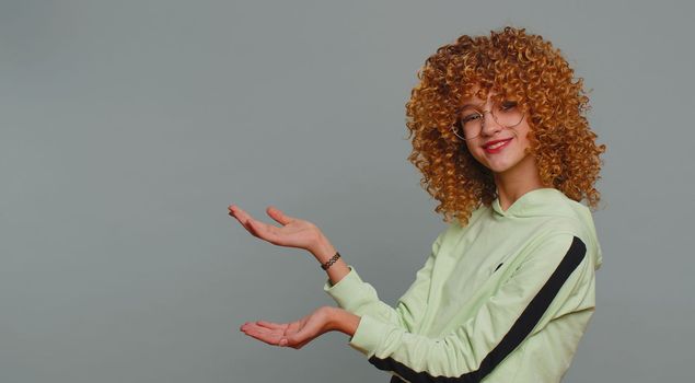 Happy teenager girl showing thumbs up pointing empty place, advertising area for commercial text copy-space for goods promotion. Young child kid with curly hair. Studio shot indoors on gray background