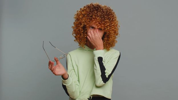 Exhausted tired teenager school girl with curly frizzy hairstyle takes off glasses, feels eyes pain, being overwork burnout from long hours studying or working. Child kid isolated on gray background