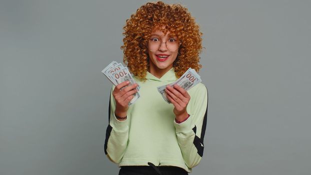 Rich pleased young teenager school girl in glasses waving money dollar cash banknotes bills like a fan, success, lottery winner, big income, wealth. Child kid with curly hair on studio gray background