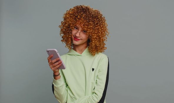 Teenager girl with curly frizzy hairstyle texting share messages content on smartphone social media applications, watching relax movie news, online store shopping sale. Child kid on gray background