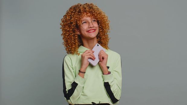 Young child girl kid with curly hair use mobile smartphone typing browsing say Wow yes found out great big win good news celebrate doing winner gesture. Teenager children on studio gray background