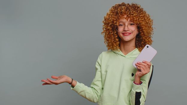 Teenager girl using smartphone pointing empty place, advertising area for commercial text copy space for goods application promotion social networks positive feedback. Child kid on gray background