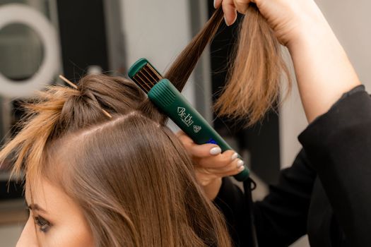 The hairdresser prepares a woman, makes a hairstyle with curls using a curling iron. Long light brown natural hair. Self care concept