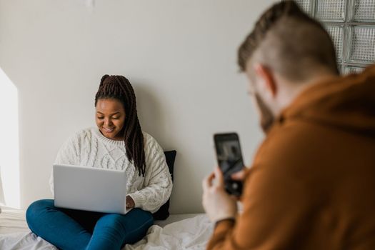 Cheerful african american woman blogger with laptop while guy making photo for blog or social media. Man taking picture girl when she writing post on social media vlog