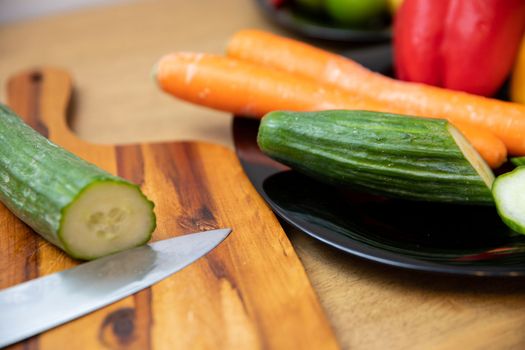 Blurred foreground, cut cucumber and knife lying on a cutting board. Next to the clear colorful vegetables laid on a black plate.
