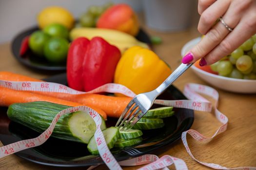 A plate with sliced cucumber and other vegetables, visible up close. A measure tape lies on top of the vegetables. A woman's hand holding a fork brings it closer to the vegetables.