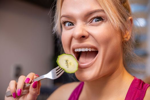 Large close-up on the face of a blonde woman with her mouth open. The woman holds a fork with a scooped cucumber slice with skin and brings it close to her open mouth.