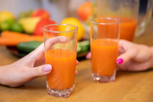 Blurred background showing plates with colorful fruits and vegetables. In the foreground, a close-up on the female hands of two people holding juice glasses.