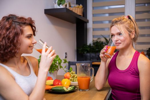 Two female friends sitting in the kitchen facing each other are drinking orange juice from tall glasses. One of the women is blonde and the other is brunette. There are plates of fruit on the table.