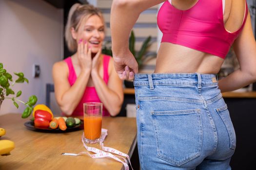 A girl standing with her back shows her friend sitting in the background the effects of weight loss by pulling back too loose pants at the waist. Women wear matching pink sports tops