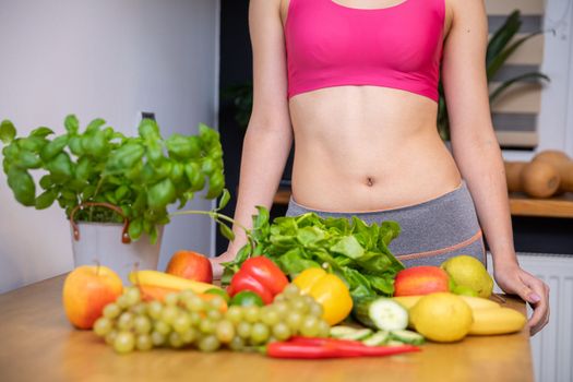 Lots of vegetables and fruits lying on the kitchen table. Behind the table a silhouette of a woman with an exposed belly in a tight pink sports top leaning against the table top
