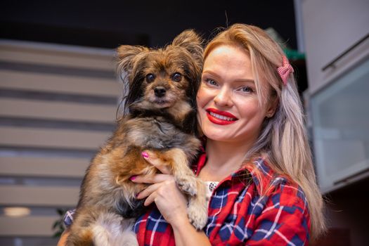 A small brown shaggy dog sitting in the arms of its owner. A woman with red lipstick cuddling a fluffy dog in her arms. A mutt from a shelter held in her arms