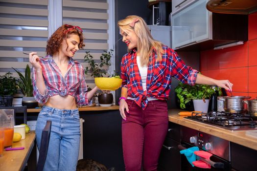 Two women dressed in checkered shirts cooking in the kitchen. The brunette holds a yellow bowl and both women look into it. The blonde woman stirs with a spoon in a metal pot on the stove.