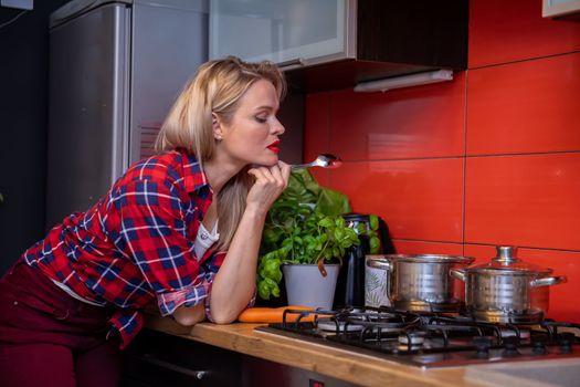 A woman dressed in a checked shirt leans her elbow against the kitchen counter and looks at a metal pot standing on a gas stove holding a spoon in her hand. Behind the stove, colorful kitchen tiles