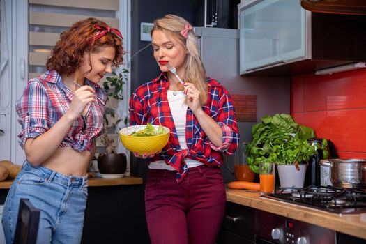 Two women standing in the kitchen eating a salad. Both women are holding forks. The blonde woman holds a yellow bowl of vegetable salad. Herbs stand on the kitchen counter. In the background is a curtained window.