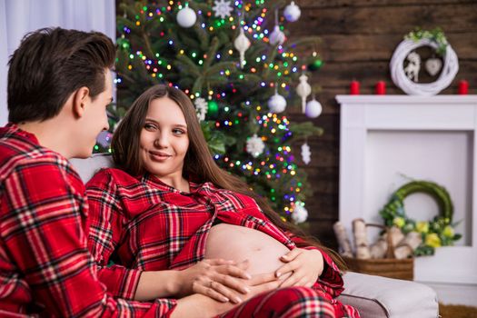 A pregnant woman sits next to her husband and smiles. In the background is a blurry dressed Christmas tree. The mother-to-be keeps her hands on her exposed pregnant belly.