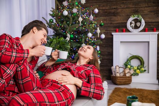 The happy parents-to-be are lying on the couch and laughing while holding white teacups, against a backdrop of a dressed Christmas tree with colorful lights. The couple is wearing the same checkered pajamas.