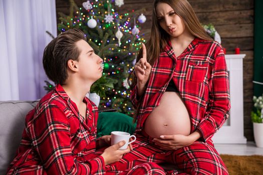 A woman exposes her large pregnant belly. The couple sits next to each other on a couch and makes funny faces at each other. A Christmas tree and other holiday decorations can be seen in the background.