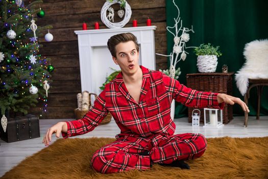 A man sits with his legs crossed on a brown carpet with long hair. He is prancing around and making funny faces, his hands raised. In the background a dressed Christmas tree and other holiday decorations