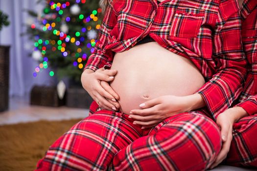 Large close-up of pregnant woman's belly holding hands on her belly. A woman's hands with an engagement ring are stroking her pregnant belly. Smudged Christmas lights in the background.