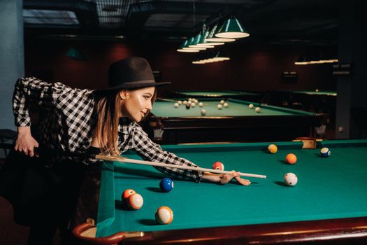 A girl in a hat in a billiard club with a cue in her hands hits a ball.Playing pool.