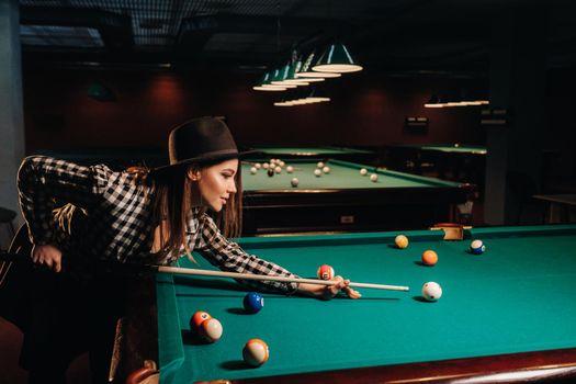 A girl in a hat in a billiard club with a cue in her hands hits a ball.Playing pool.