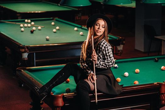 A girl in a hat in a billiard club sits on a billiard table with a cue in her hands.Playing billiards.
