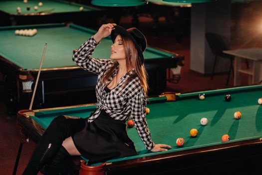 A girl in a hat in a billiard club sits on a billiard table.Playing pool.