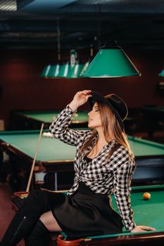 A girl in a hat in a billiard club sits on a billiard table.Playing pool.