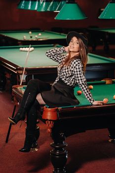 A girl in a hat in a billiard club sits on a billiard table.Playing pool.