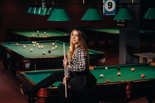 A girl in a hat in a billiard club sits on a billiard table with a cue in her hands.Playing billiards.