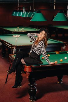 A girl in a hat in a billiard club sits on a billiard table with a cue in her hands.Playing billiards.