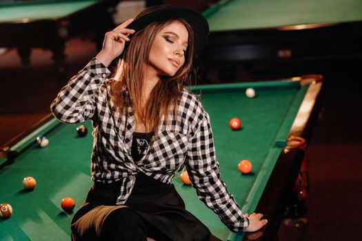 A girl in a hat in a billiard club sits on a billiard table.Playing pool.