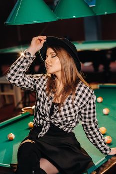 A girl in a hat in a billiard club sits on a billiard table.Playing pool.