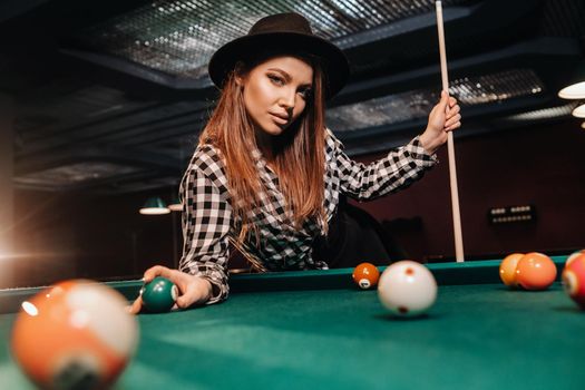 A girl in a hat in a billiard club with a cue and balls in her hands.Playing pool.