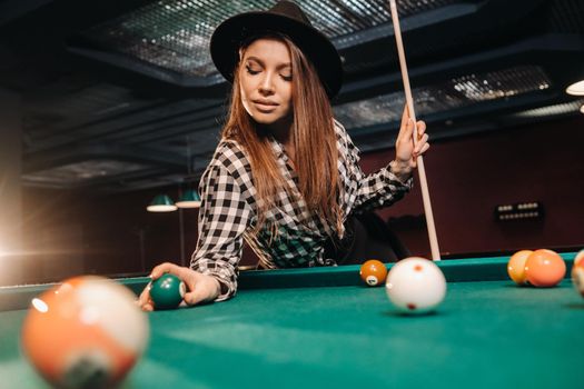 A girl in a hat in a billiard club with a cue and balls in her hands.Playing pool.