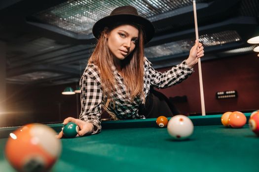 A girl in a hat in a billiard club with a cue and balls in her hands.Playing pool.