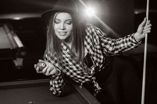 A girl in a hat in a billiard club with a cue and balls in her hands.Playing pool. black and white photo.