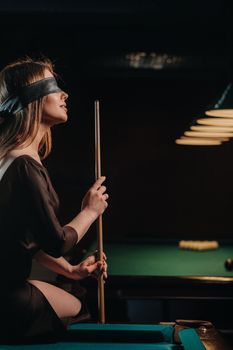 A girl with a blindfold and a cue in her hands is sitting on a table in a billiard club.Russian billiards.