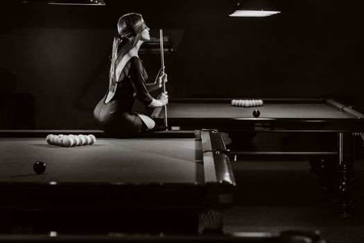 A girl with a blindfold and a cue in her hands is sitting on a table in a billiard club.Russian billiards. black and white photo.