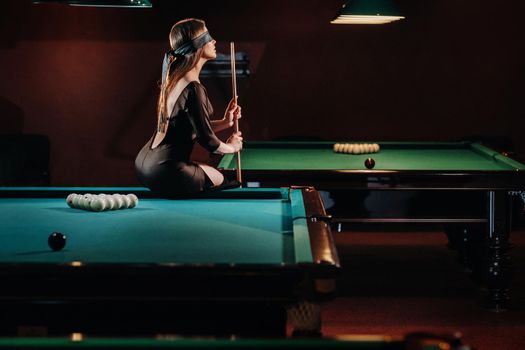 A girl with a blindfold and a cue in her hands is sitting on a table in a billiard club.Russian billiards.