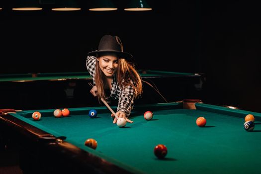 A girl in a hat in a billiard club with a cue in her hands hits a ball.Playing billiards.