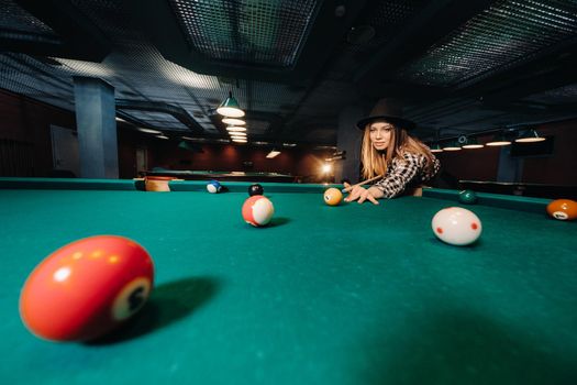A girl in a hat in a billiard club with a cue in her hands hits a ball.Playing pool.
